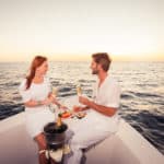 happy smiling couple sitting on bow of motor boat celebrating with champagne and fingerfood on sea at sunset