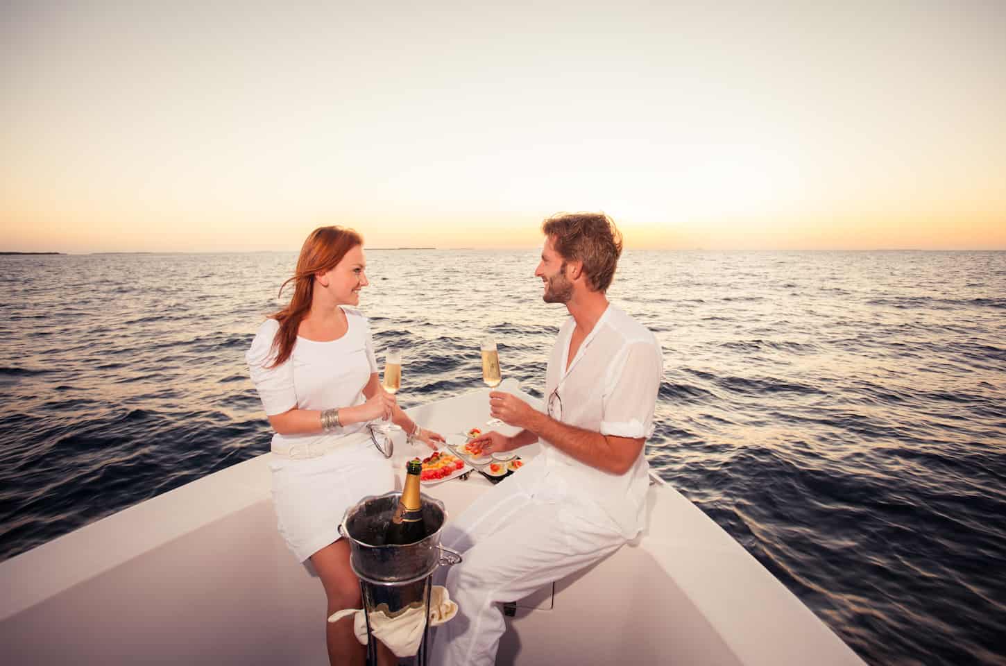 happy smiling couple sitting on bow of motor boat celebrating with champagne and fingerfood on sea at sunset